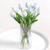 A bouquet of white and light blue artificial tulip flowers in a clear glass vase on a white plate.