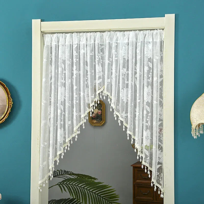White embroidered sheer lace door curtain with tassels hanging in a white frame against a teal wall.