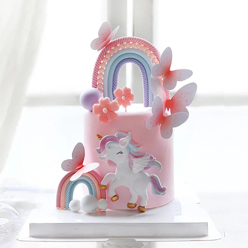 A pink unicorn theme cake topper set featuring a rainbow arch and pink butterfly decorations on a cake.