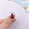 A small, red and black resin beetle rhinestone applique is held between a thumb and finger.
