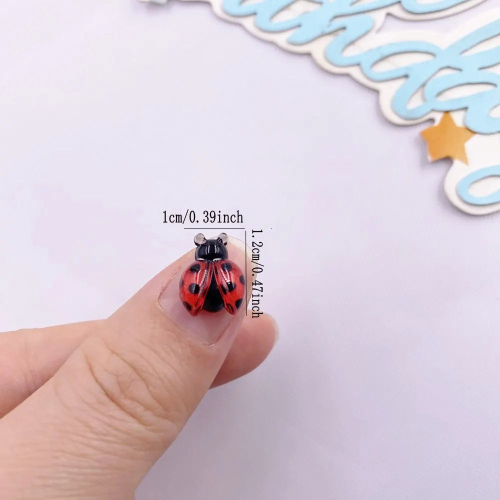 A small, red and black resin beetle rhinestone applique is held between a thumb and finger.
