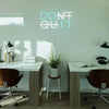 A glowing neon sign spelling DON'T QUIT hangs above a modern desk with plants and office equipment.