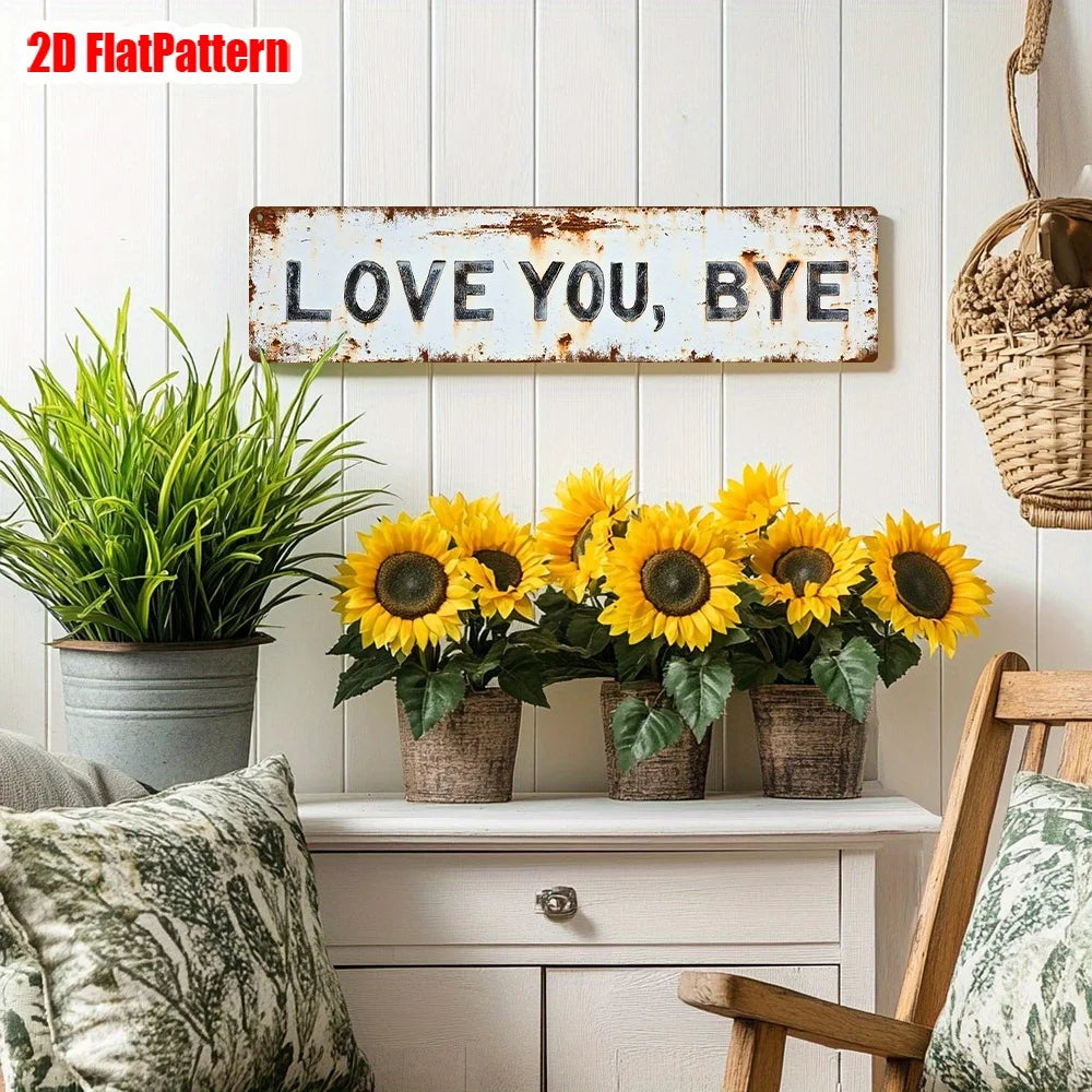 A rustic wooden sign with the text LOVE YOU BYE mounted on a white wall, surrounded by potted sunflowers and greenery.