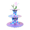 A three-tier rainbow cupcake stand featuring purple, pink, and blue iridescent layers with a green figure on top.