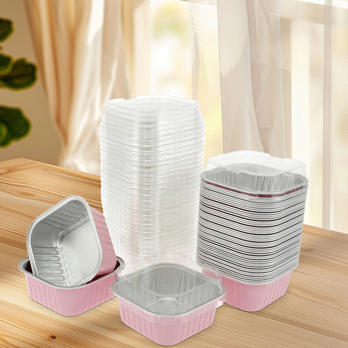 A stack of clear reusable aluminum foil baking cups with lids and spoons in pink and silver on a wooden surface.