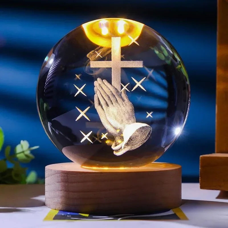 A glass crystal ball night light with a glowing yellow light, featuring a golden cross and a hand praying inside.