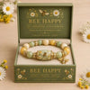 A gold and pearl stretch charm bracelet set in a green box featuring bee motifs and the text Bee Happy