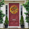 A red door decorated with an artificial flower wreath and potted evergreen trees.
