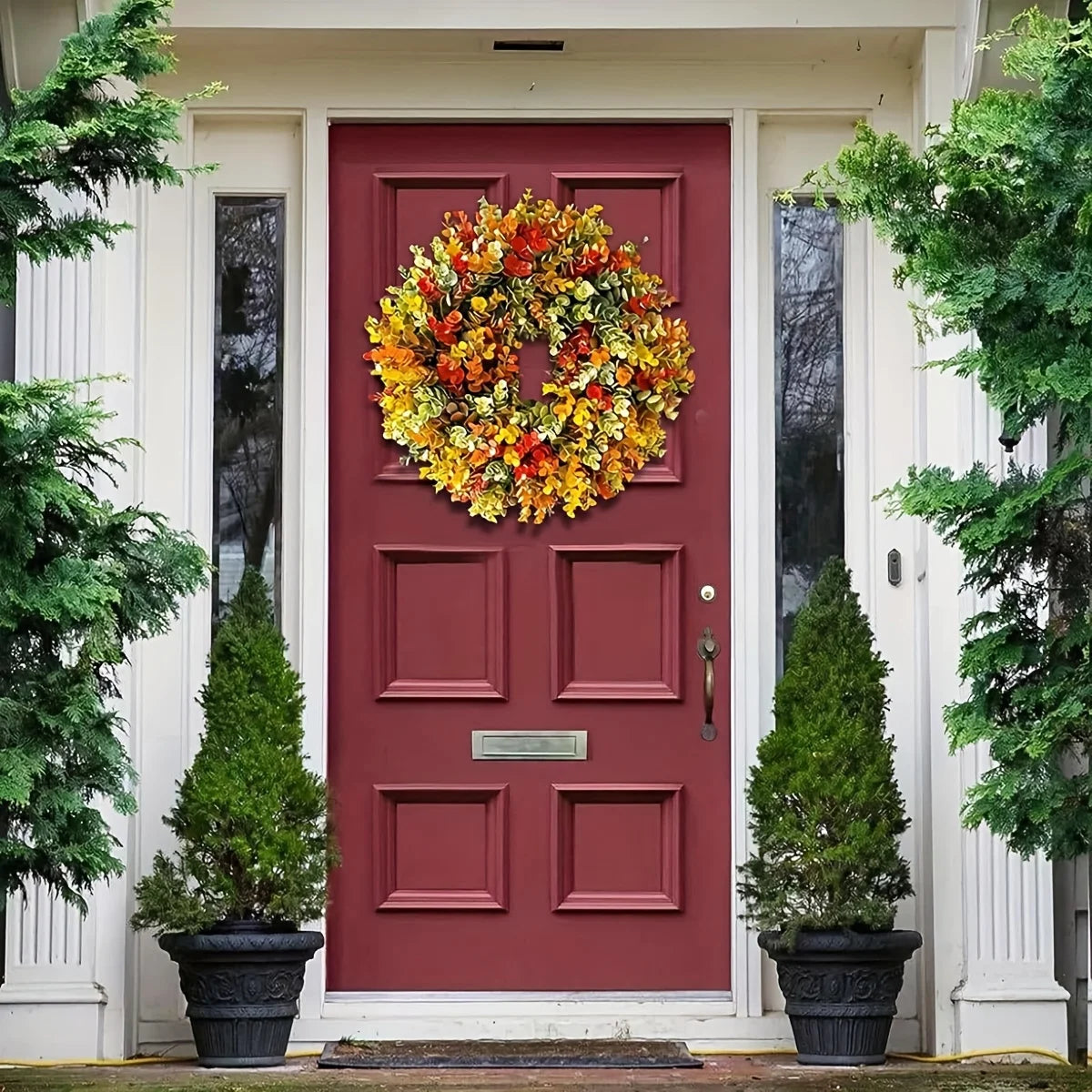 A red door decorated with an artificial flower wreath and potted evergreen trees.