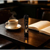 A dark wooden table features a cup of coffee, an open book, and a black 3D printed wishing willow stick candle.