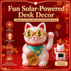 A white cat ornament wearing gold glasses and a green collar sits on a red base, decorated with coins, next to a solar panel.