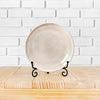 A round, matte beige ceramic plate displayed on a black metal easel against a white brick background.