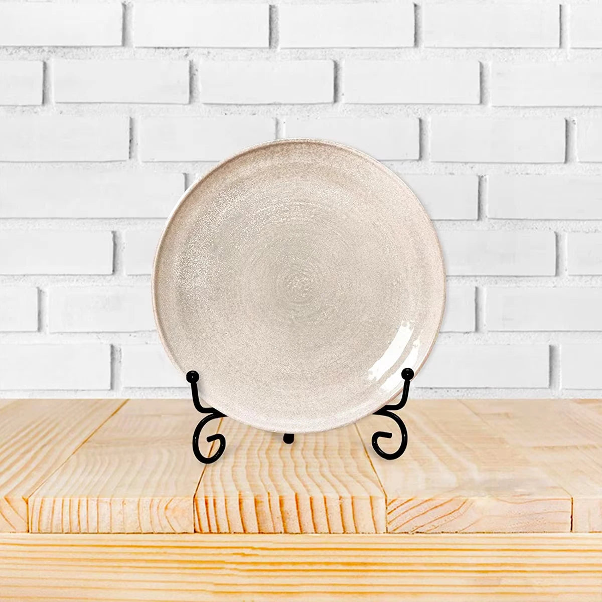 A round, matte beige ceramic plate displayed on a black metal easel against a white brick background.