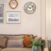 A rustic wooden wall clock with a blue and white face hangs on a light-colored wall above a sofa.