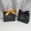 Two black paperboard thank you gift bags tied with gold ribbon are displayed on a white surface.