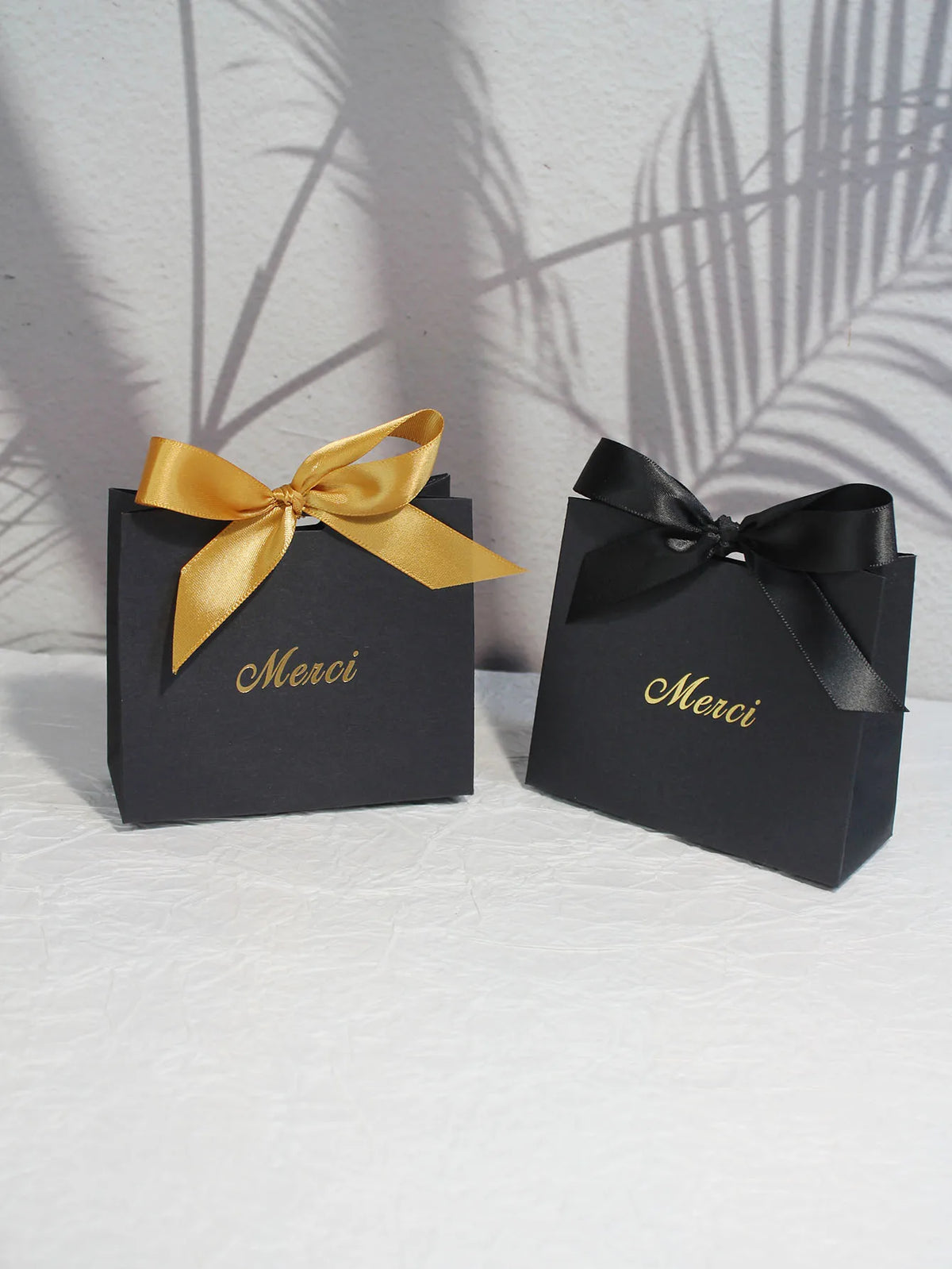 Two black paperboard thank you gift bags tied with gold ribbon are displayed on a white surface.
