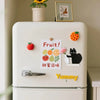 A white refrigerator decorated with fruit and a black cat theme featuring magnetic acrylic decor.