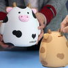 A white and black cow-shaped piggy bank is being held by a person, next to an orange cow piggy bank.