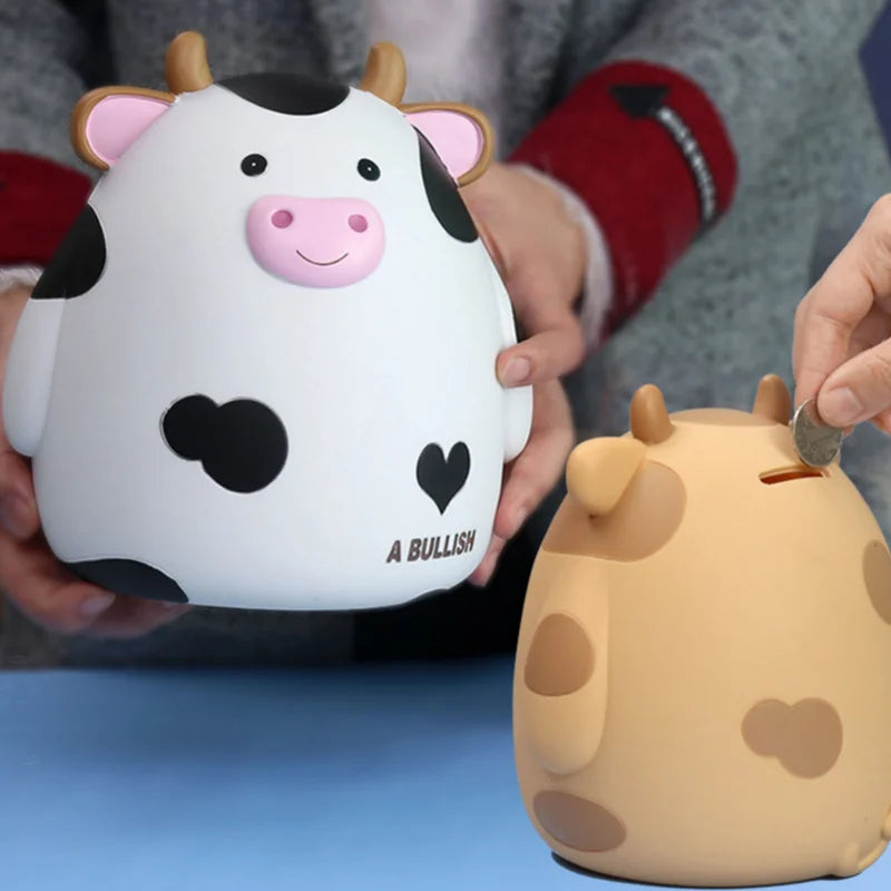 A white and black cow-shaped piggy bank is being held by a person, next to an orange cow piggy bank.
