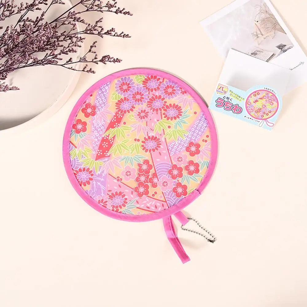 A pink portable foldable cooling handheld fan with a floral pattern is shown next to some paper and a branch of dried flowers