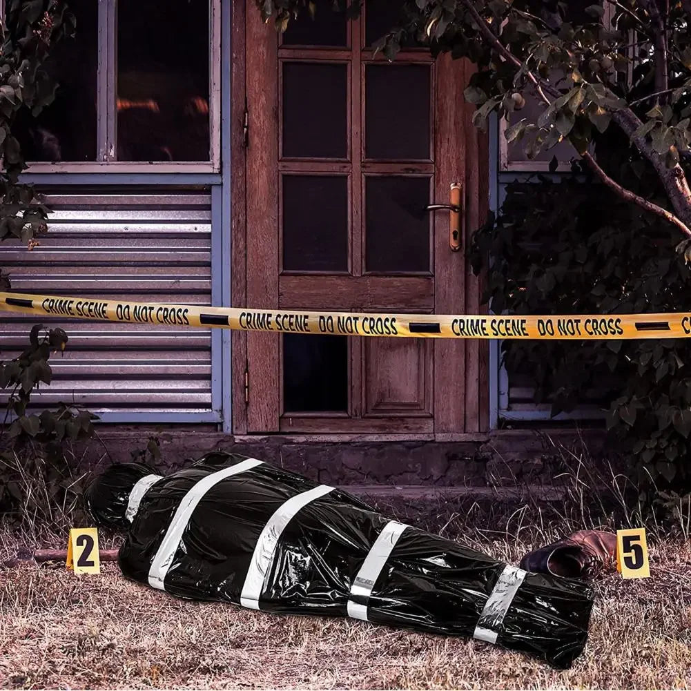 A realistic dead victim prop covered in black plastic and white stripes lying on grass near a wooden door.