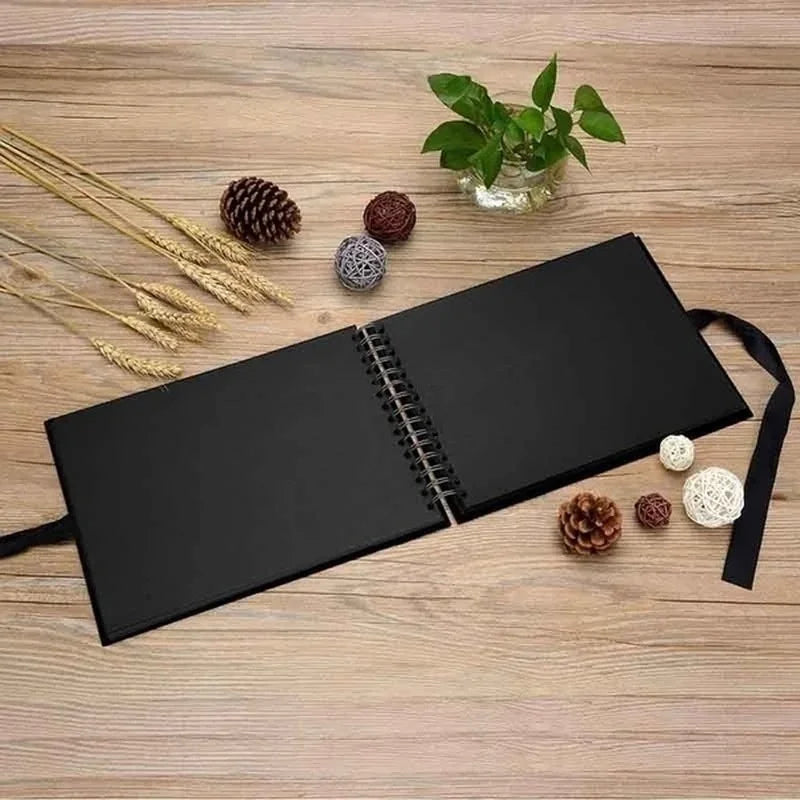 A black DIY scrapbook photo album lying on a wooden surface with pinecones and dried grasses.