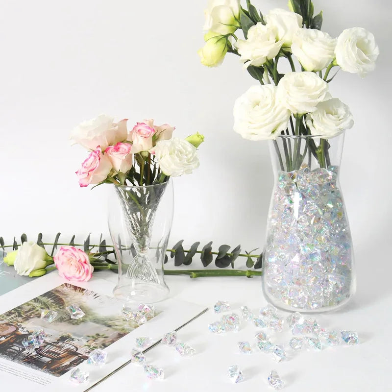 A glass vase holds white roses and green foliage, with crushed diamond acrylic pebbles scattered on the surface.