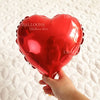 A hand holds a shiny red heart foil balloon against a white quilted background.