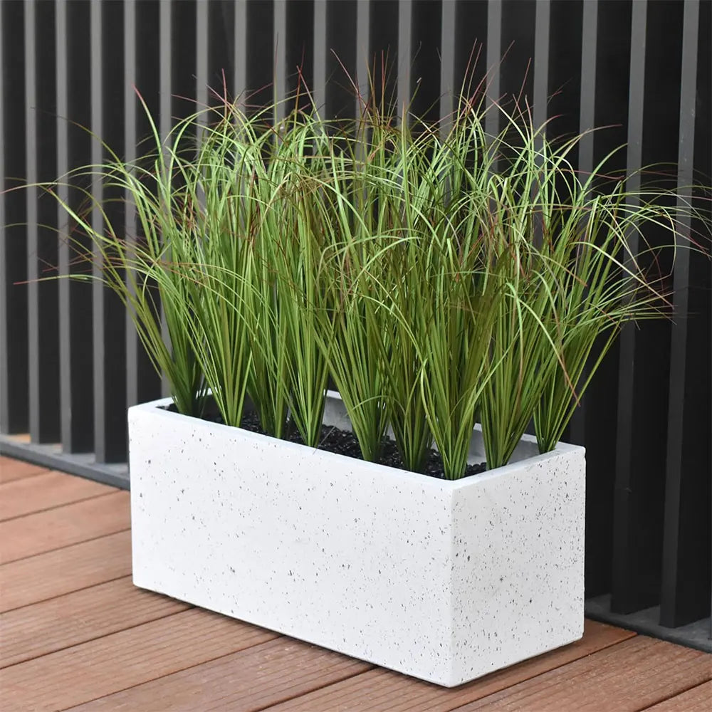 A white planter filled with artificial grass greenery bundle set against a dark vertical slat background.