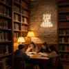 A group of students studying at a wooden table in a library setting with a neon sign on the stone wall.