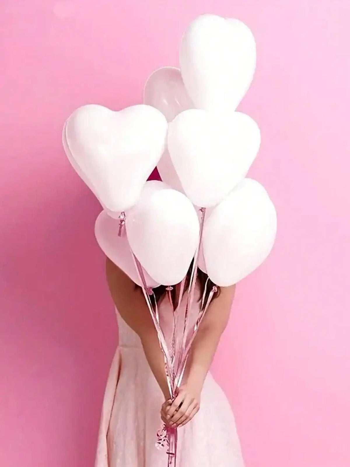 A person holds a bouquet of white heart-shaped latex balloons against a pink background.