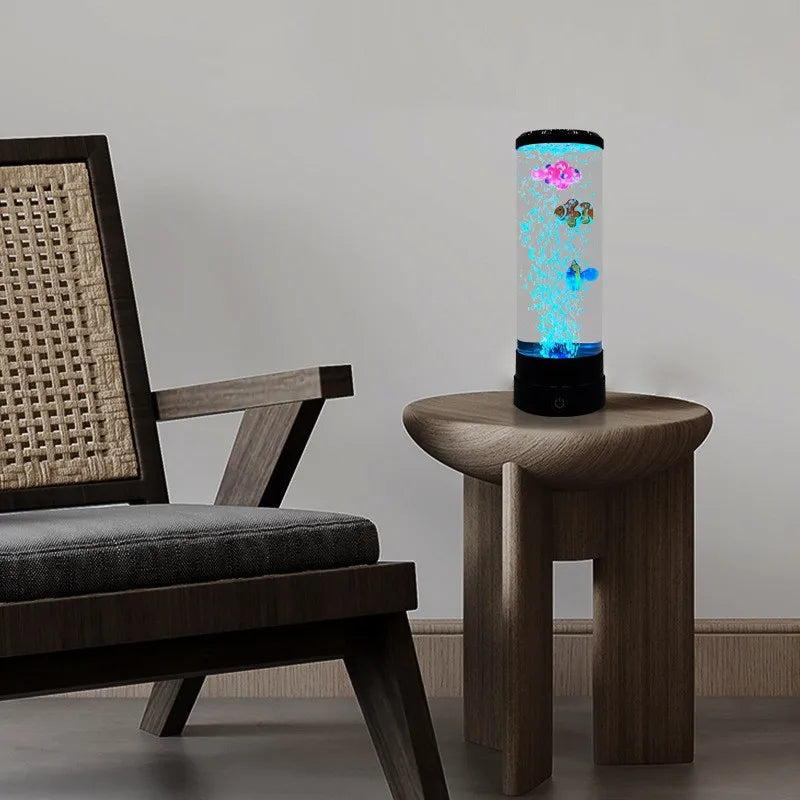 A tall, cylindrical LED fish lamp with glowing blue and pink lights sits on a wooden table next to a dark chair.