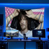 A man with long dark hair is holding a large American flag banner in front of him, set against a blue background.
