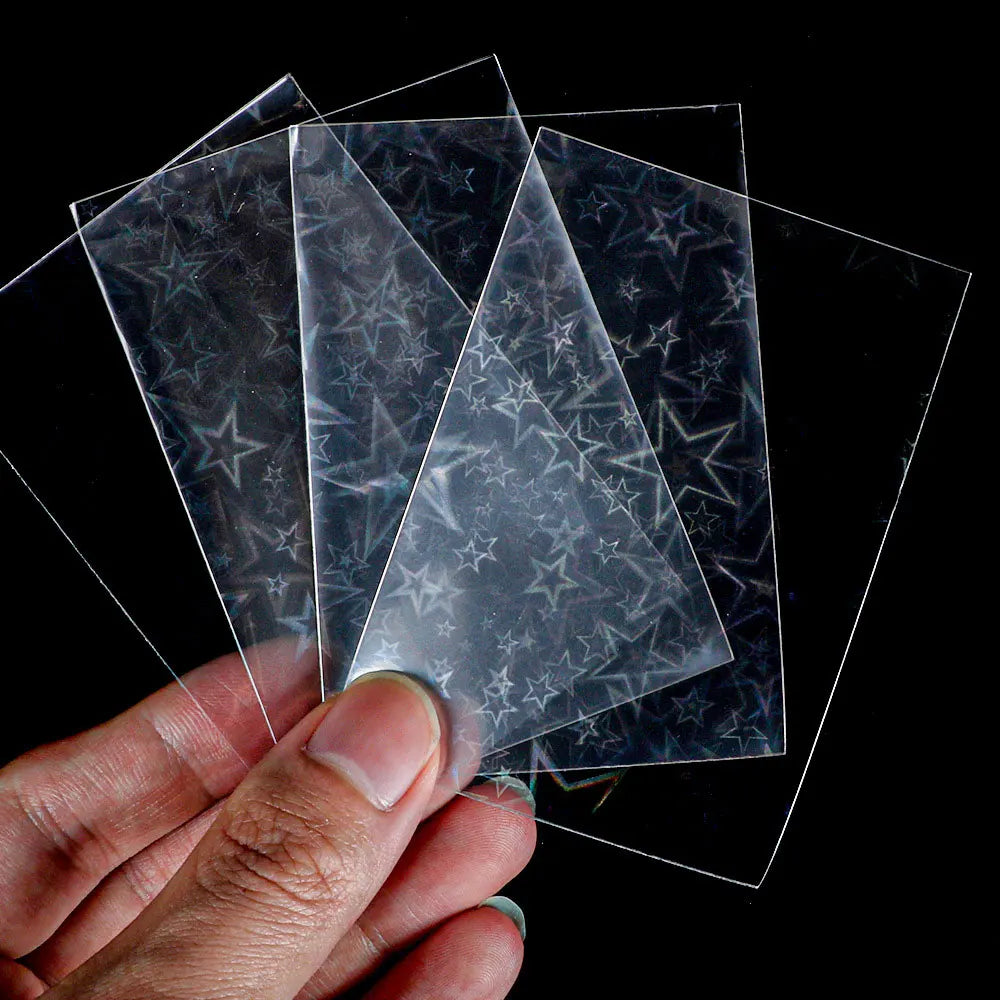 A hand holds several holographic card sleeves, clear plastic protectors with star patterns, against a black background.
