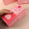 A hand holds a stack of pink pop-up surprise gift boxes with heart designs on a light background.