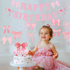 A baby girl in a pink dress sits in front of a cake and a glitter pink bow birthday banner.