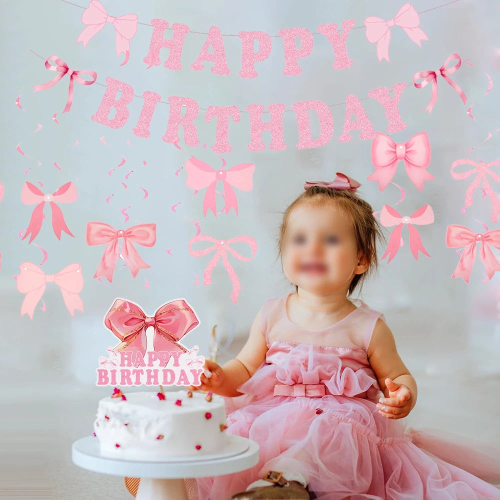 A baby girl in a pink dress sits in front of a cake and a glitter pink bow birthday banner.