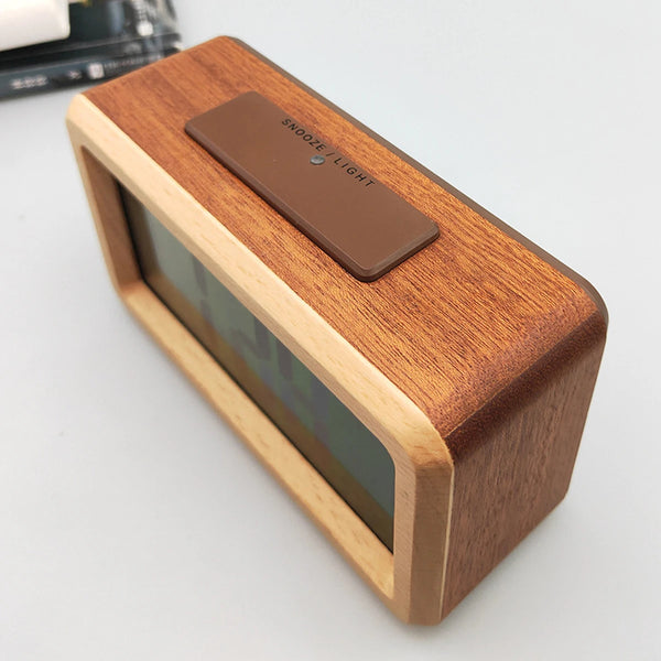 A wooden digital alarm clock with a dark brown face and light wood casing is shown on a white surface.