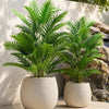 A potted artificial areca palm tree with vibrant green fronds set against a stone wall backdrop.
