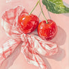 A watercolor painting of two bright red cherries tied with a pink and white checkered ribbon on a light pink background.
