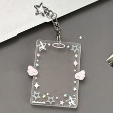 A clear acrylic photocard holder with stars, lines, and wings is attached to a silver keychain.