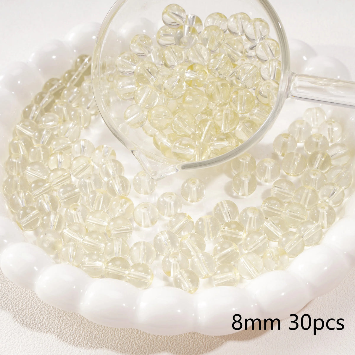 A clear glass measuring spoon holds small round beads scattered on a white dish.