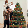 A woman and a man decorate a large Christmas tree with festive red and green tinsel garland.
