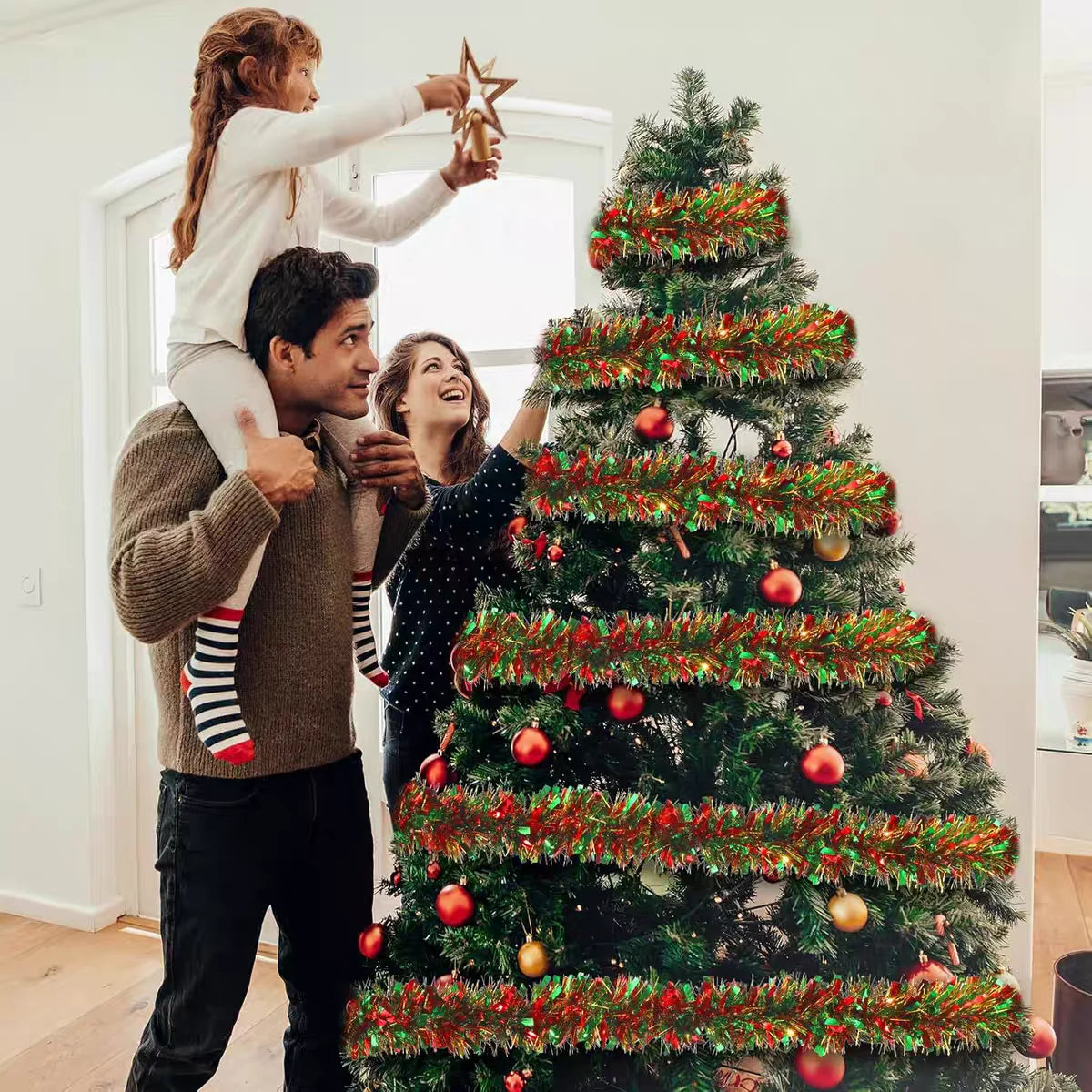 A woman and a man decorate a large Christmas tree with festive red and green tinsel garland.