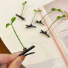 A hand holds a black bean sprout hair clip with a grass top, showing several similar clips on a white surface.