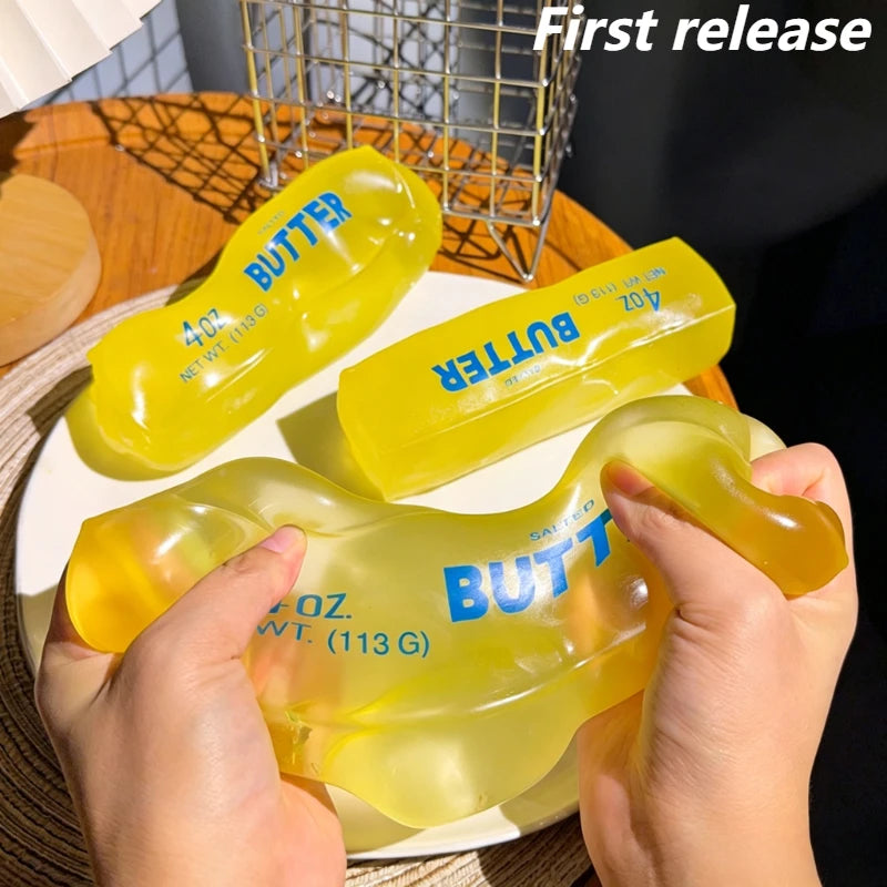 A hand holds a yellow butter squeeze toy, labeled 4 oz. net wt. (113 g), against a background of a wire basket.