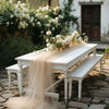 A white wooden table set outdoors with a sheer beige tulle fabric draped over it, surrounded by greenery and white flowers.
