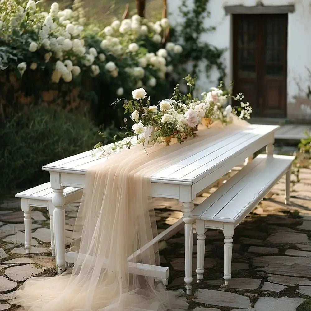 A white wooden table set outdoors with a sheer beige tulle fabric draped over it, surrounded by greenery and white flowers.