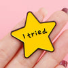 A hand holds a yellow star enamel pin with the text I tried on a pink background.