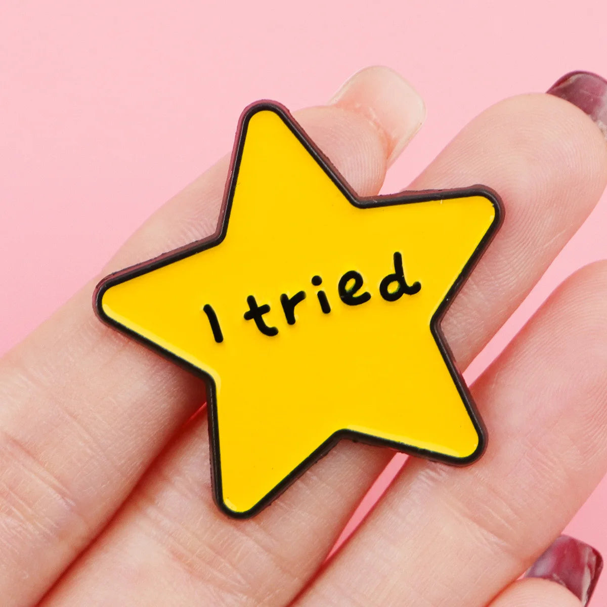 A hand holds a yellow star enamel pin with the text I tried on a pink background.